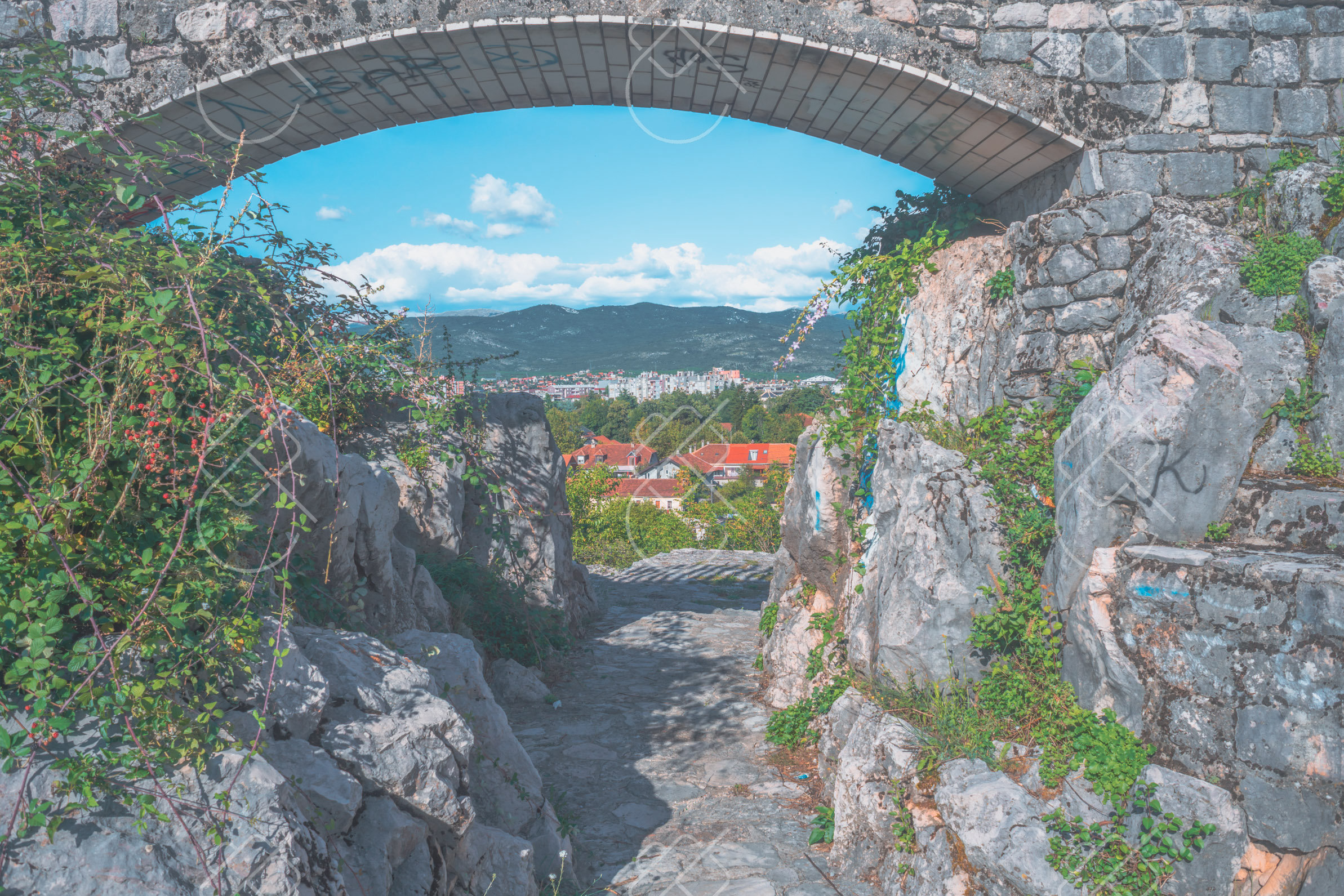 A stone arch over the rocky approach to the fortress, framing Nikšić and the hills beyond — Bedem Fortress, fine art travel photography print available from Clever Tourists
